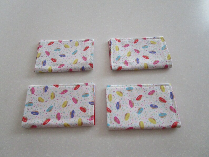 May include: Four white fabric napkins with a donut and sprinkles pattern. The donuts are pink, yellow, blue, and green.