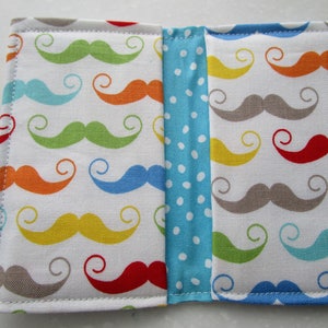 May include: A white fabric wallet with a colorful mustache pattern. The wallet is divided into two sections with a blue and white polka dot fabric strip in the middle.