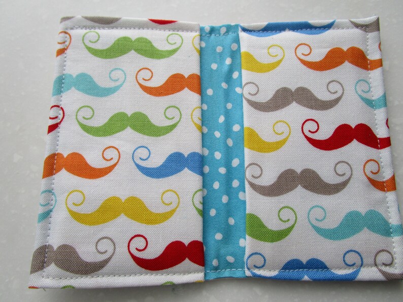 May include: A white fabric wallet with a colorful mustache pattern. The wallet is divided into two sections with a blue and white polka dot fabric strip in the middle.