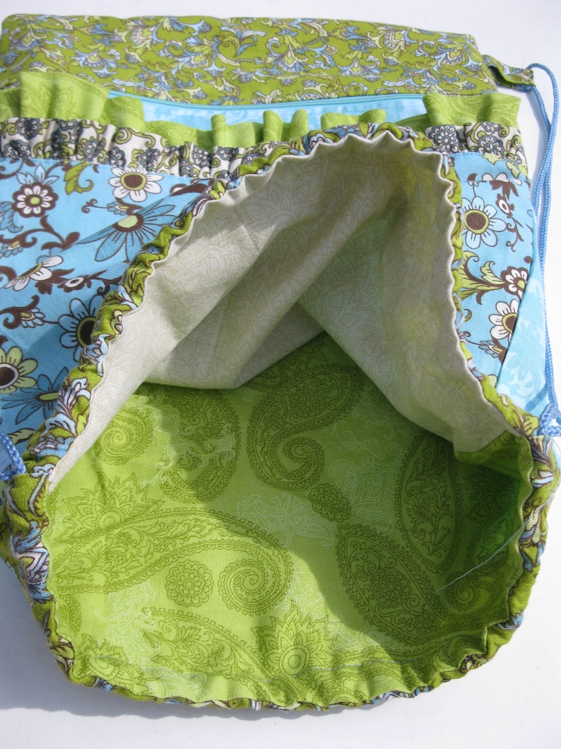 May include: A green and blue fabric drawstring bag with a paisley pattern lining. The bag has a ruffled trim and a zippered pocket.