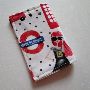 Puede incluir: Una bolsa de tela blanca con un dise&ntilde;o de metro de Londres rojo, blanco y azul. La bolsa presenta un c&iacute;rculo rojo con las palabras "UNDERGROUND" en blanco, una cabina telef&oacute;nica roja y una mujer con cabello rojo que lleva gafas de sol y un vestido negro.