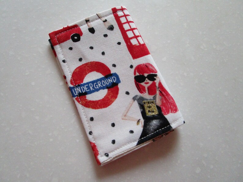Puede incluir: Una bolsa de tela blanca con un dise&ntilde;o de metro de Londres rojo, blanco y azul. La bolsa presenta un c&iacute;rculo rojo con las palabras "UNDERGROUND" en blanco, una cabina telef&oacute;nica roja y una mujer con cabello rojo que lleva gafas de sol y un vestido negro.