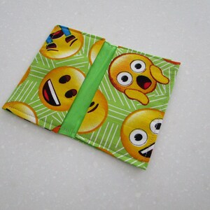 May include: A square, fabric wallet featuring a repeating pattern of yellow emoji faces on a light green background with white lines. A vertical green stripe accents the center. The wallet is closed.