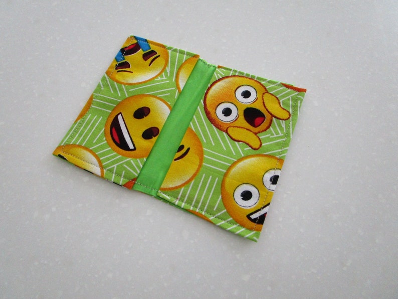 May include: A square, fabric wallet featuring a repeating pattern of yellow emoji faces on a light green background with white lines. A vertical green stripe accents the center. The wallet is closed.