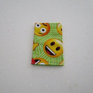 May include: A rectangular, folded fabric item featuring a repeating pattern of yellow emoji faces on a green background with white diagonal lines. The emojis display various expressions, including smiling and winking.