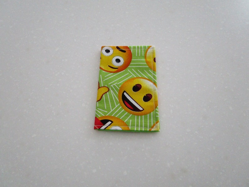 May include: A rectangular, folded fabric item featuring a repeating pattern of yellow emoji faces on a green background with white diagonal lines. The emojis display various expressions, including smiling and winking.