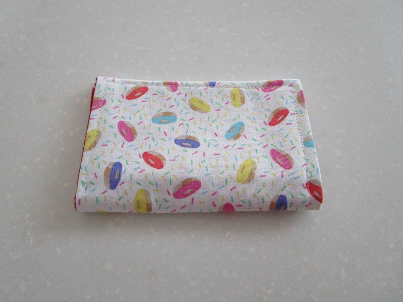 May include: A white fabric pouch with a red border and a pattern of colorful donuts and sprinkles.