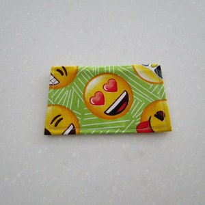 May include: A small, rectangular fabric item featuring a repeating pattern of yellow emoji faces with various expressions, including hearts for eyes and a tongue sticking out. The background is light green with white diagonal lines.