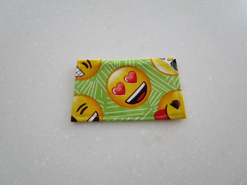 May include: A small, rectangular fabric item featuring a repeating pattern of yellow emoji faces with various expressions, including hearts for eyes and a tongue sticking out. The background is light green with white diagonal lines.