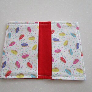 May include: A white fabric book cover with a red fabric spine. The front and back covers feature a pattern of colorful donuts and sprinkles.