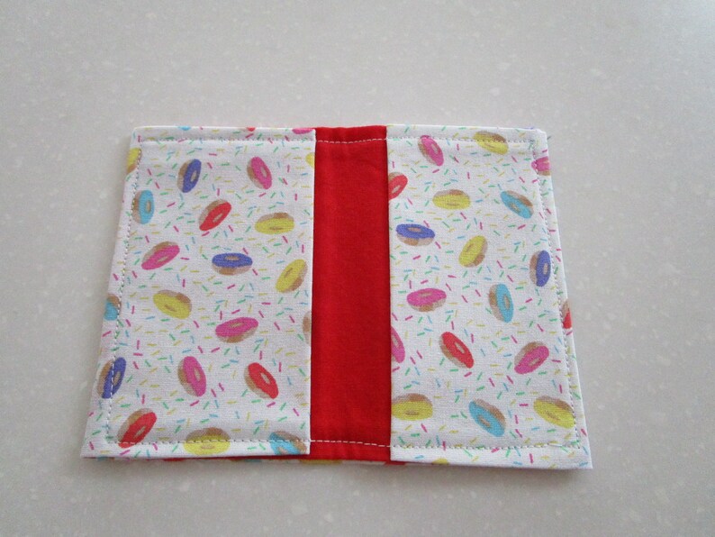 May include: A white fabric book cover with a red fabric spine. The front and back covers feature a pattern of colorful donuts and sprinkles.