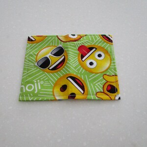 May include: A square, green Emoji-themed fabric coaster featuring various yellow emoji faces with sunglasses, tongues out, and other expressions. The word "moji" is printed in white on the bottom left corner. The coaster is likely made of cotton or a similar fabric.