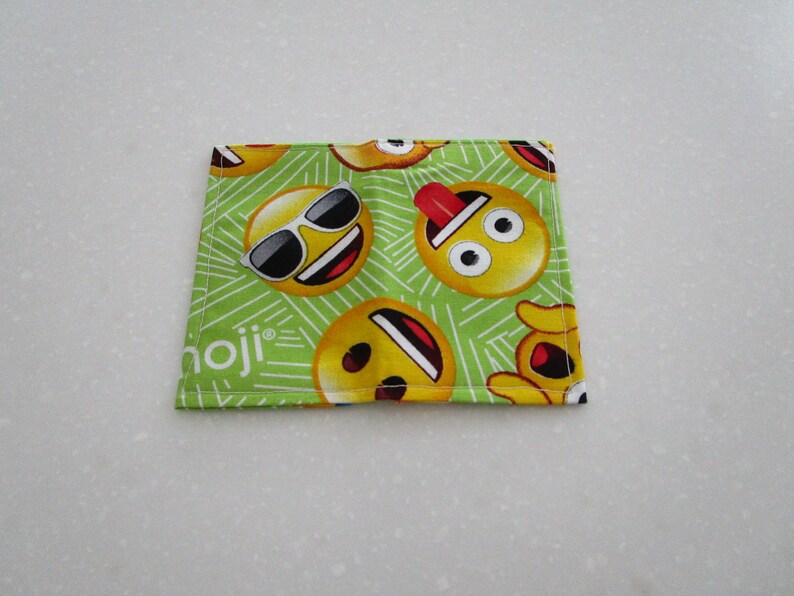 May include: A square, green Emoji-themed fabric coaster featuring various yellow emoji faces with sunglasses, tongues out, and other expressions. The word "moji" is printed in white on the bottom left corner. The coaster is likely made of cotton or a similar fabric.