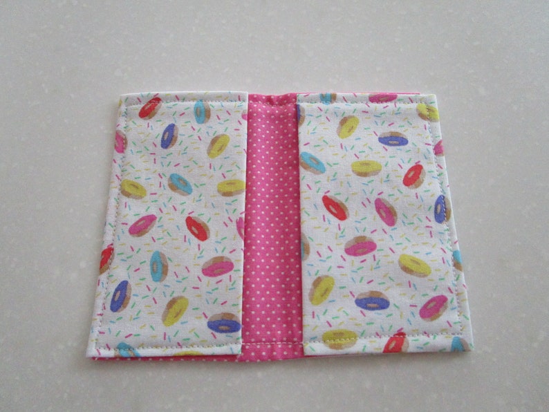 May include: A pink and white fabric wallet with a donut and sprinkles pattern. The wallet has two sections and is closed with a zipper.