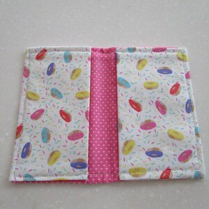 May include: A pink and white fabric wallet with a donut and sprinkles pattern. The wallet has two sections and is closed with a zipper.