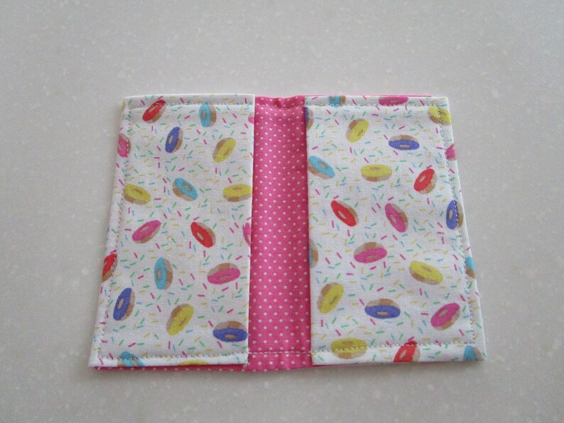 May include: A pink and white fabric wallet with a donut and sprinkles pattern. The wallet has two sections and is closed with a zipper.