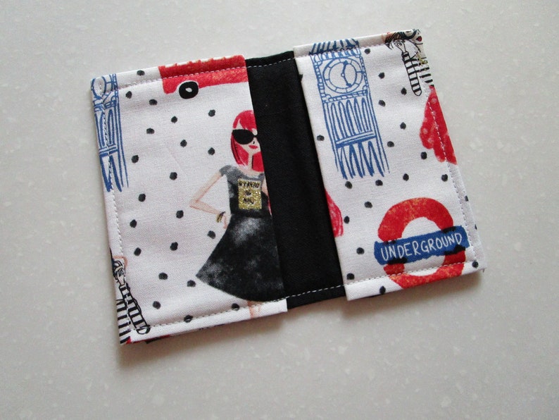 Puede incluir: Una cartera de tela blanca con forro de tela negro. La parte delantera de la cartera presenta una ilustraci&oacute;n en blanco y negro de una mujer con un vestido negro y gafas de sol. La parte posterior de la cartera presenta una ilustraci&oacute;n roja y azul del Big Ben y el logotipo del metro de Londres.