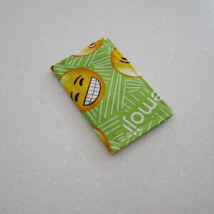 May include: A rectangular, folded fabric item featuring a repeating pattern of yellow smiling emoji faces on a bright green background. The word "emoji" is printed in white along one edge. White lines create a striped design.