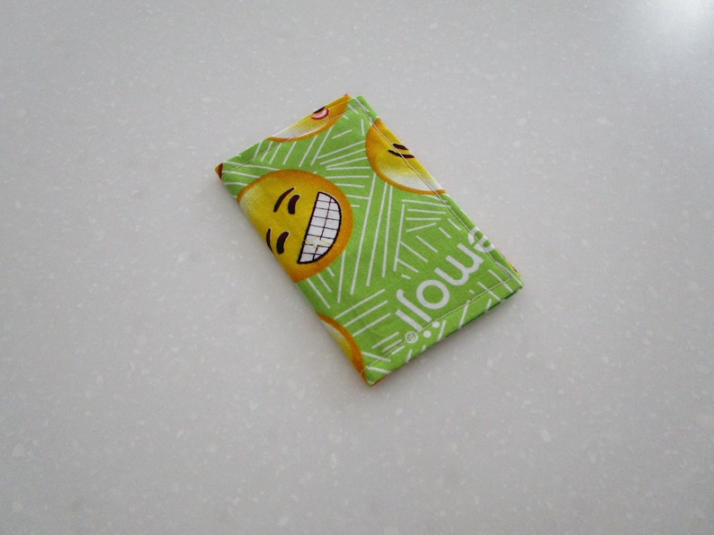 May include: A rectangular, folded fabric item featuring a repeating pattern of yellow smiling emoji faces on a bright green background. The word "emoji" is printed in white along one edge. White lines create a striped design.