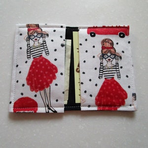 Puede incluir: Una cartera de tela blanca con un patr&oacute;n de lunares negros. La cartera presenta una ilustraci&oacute;n roja y blanca de una mujer con gafas y una falda roja. La cartera tiene una solapa que se cierra con un broche a presi&oacute;n.