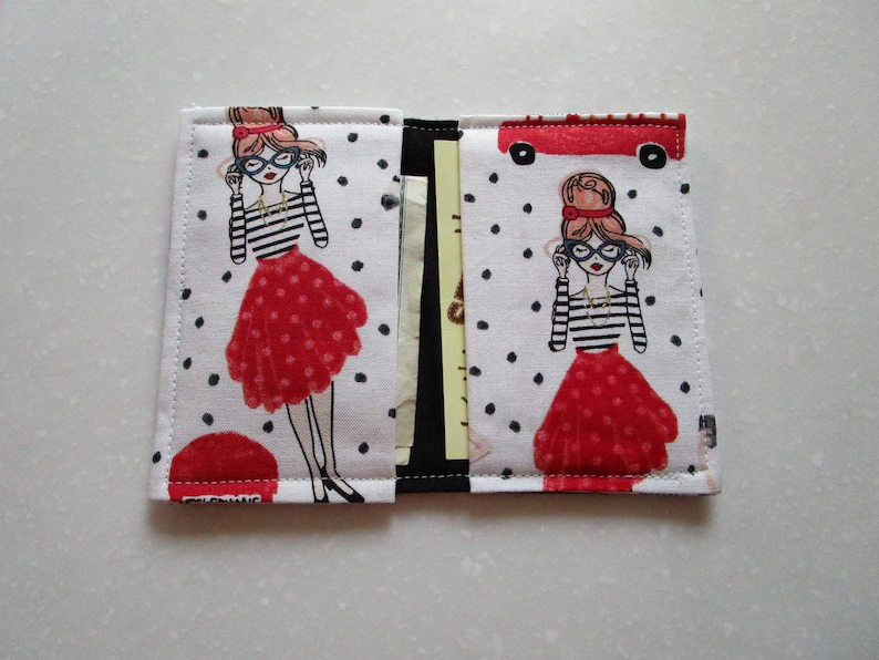 Puede incluir: Una cartera de tela blanca con un patr&oacute;n de lunares negros. La cartera presenta una ilustraci&oacute;n roja y blanca de una mujer con gafas y una falda roja. La cartera tiene una solapa que se cierra con un broche a presi&oacute;n.