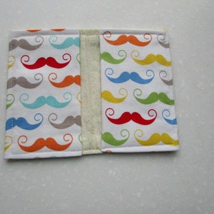 May include: A white fabric book cover with a colorful pattern of mustaches. The mustaches are in various colors, including red, yellow, blue, green, and gray.
