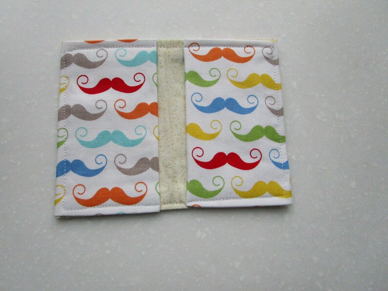 May include: A white fabric book cover with a colorful pattern of mustaches. The mustaches are in various colors, including red, yellow, blue, green, and gray.