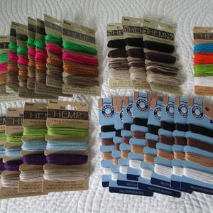 May include: A collection of hemp cord spools in various colors, including green, pink, brown, black, and blue. The spools are neatly arranged on a white surface, with each card labeled with the word "HEMP".