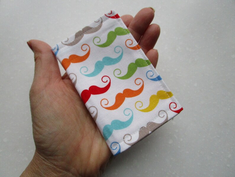 May include: A small white fabric pouch with a colorful mustache pattern. The pouch is made of cotton fabric and has a beige seam around the edges.