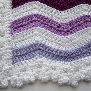 May include: A crocheted blanket with a wavy pattern in shades of purple, lavender, and white. The blanket has a scalloped edge.