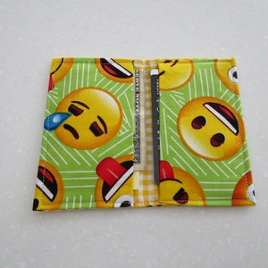 May include: A fabric wallet with a green background and a pattern of yellow emoji faces. The wallet is open, revealing a credit card and a dollar bill. The emoji faces have various expressions.