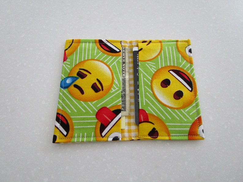 May include: A fabric wallet with a green background and a pattern of yellow emoji faces. The wallet is open, revealing a credit card and a dollar bill. The emoji faces have various expressions.