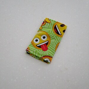 May include: A folded, rectangular cloth with a repeating pattern of yellow emoji faces on a green background. The emojis have various expressions, including smiling faces with tongues sticking out. The cloth is likely a handkerchief or small towel.