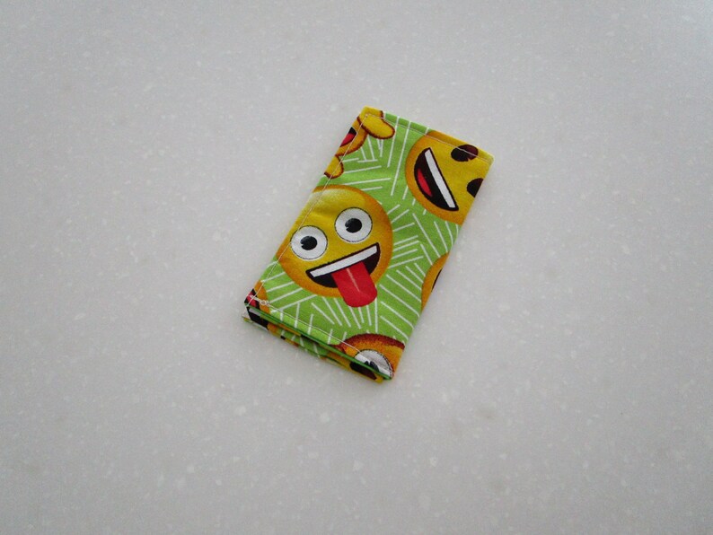 May include: A folded, rectangular cloth with a repeating pattern of yellow emoji faces on a green background. The emojis have various expressions, including smiling faces with tongues sticking out. The cloth is likely a handkerchief or small towel.