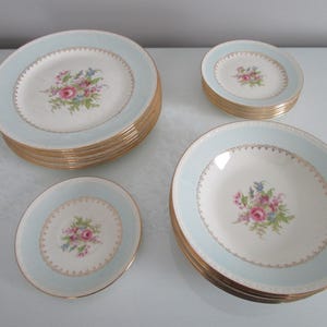 May include: A collection of vintage porcelain dishware. The set includes dinner plates, salad plates, and bowls. Each piece features a light blue border, gold trim, and a floral bouquet design in the center. The plates are stacked neatly.