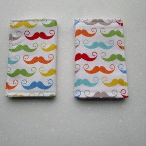 May include: Two fabric passport holders with a colorful mustache pattern. The mustaches are in shades of red, orange, yellow, green, blue, and gray.