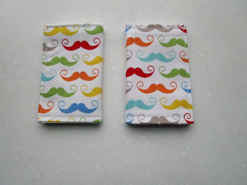 May include: Two fabric passport holders with a colorful mustache pattern. The mustaches are in shades of red, orange, yellow, green, blue, and gray.