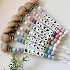 May include: A set of ten personalized silicone baby teethers with wooden clips. Each teether features a different name spelled out in white beads with black lettering. The teethers are in various colors, including pink, purple, blue, green, and beige.