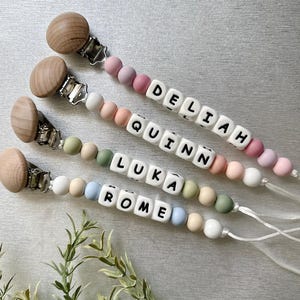 May include: Four silicone baby teethers with wooden clips and personalized name beads. The beads are in pastel colors and spell out the names "DeLiah", "Quinn", "Luka", and "Rome".