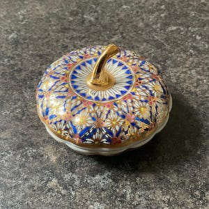 May include: A ceramic jar with a lid, decorated with a floral pattern in white, blue, pink, and gold. The jar has a gold stem-like handle and a scalloped edge. The jar is round and sits on a grey surface.