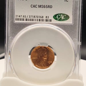 May include: A 1952-D Lincoln cent coin, graded CAC MS65RD, encased in a clear protective holder. The coin features a portrait and the inscription "IN GOD WE TRUST". The holder displays the grade and certification details.