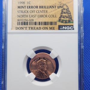 May include: A 1998 1C penny coin, graded and encased in a protective holder. The coin is off-center and features the text "DON'T TREAD ON ME". The holder includes a label with details about the coin's error and grading.