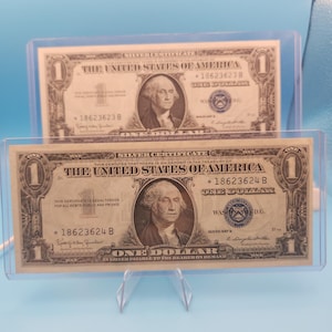 May include: Two United States one-dollar silver certificates encased in clear plastic holders. Each banknote features a portrait of George Washington, the text "The United States of America," "One Dollar," and serial numbers 18623623B and 18623624B.