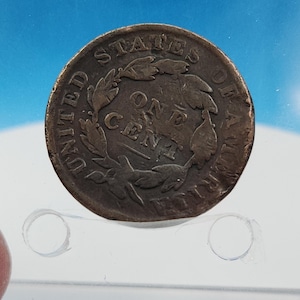 May include: A copper United States One Cent coin. The coin features the words "UNITED STATES OF AMERICA" and "ONE CENT" with a wreath design. The coin is a dark brown color and appears to be aged.