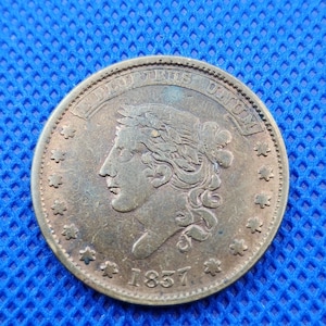 May include: A round copper coin featuring a profile of Liberty facing left, surrounded by thirteen stars. The text E PLURIBUS UNUM is arched above the head, and the year 1857 is inscribed at the bottom. The coin has a metallic, weathered finish.