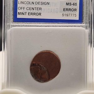 May include: A copper Lincoln penny with an off-center mint error, encapsulated in a clear plastic holder. The holder has a blue label with text including "LINCOLN DESIGN" and "MINT ERROR". The coin is graded MS-65.
