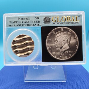 May include: A rectangular plastic slab containing a Kennedy 50C coin, labeled Waffle Cancelled Brilliant Uncirculated by Global Certification Services, Inc. The slab holds a metallic, wavy-textured circular piece and a 2003 Kennedy half dollar.