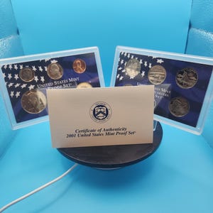 May include: A 2001 United States Mint Proof Set, featuring various coins in protective cases. The set includes a Certificate of Authenticity. The coins are displayed on a black circular stand against a blue backdrop.