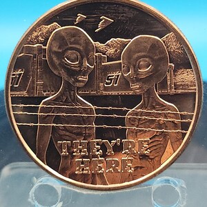 May include: A bronze-colored coin featuring two alien figures facing each other, with the text "THEY'RE HERE" below. The background includes a depiction of a fence, mountains, and the number "51".