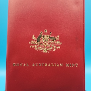 May include: A red leather-bound case with gold lettering and a gold emblem. The text "ROYAL AUSTRALIAN MINT" is displayed on the front. The emblem features a kangaroo and an emu.